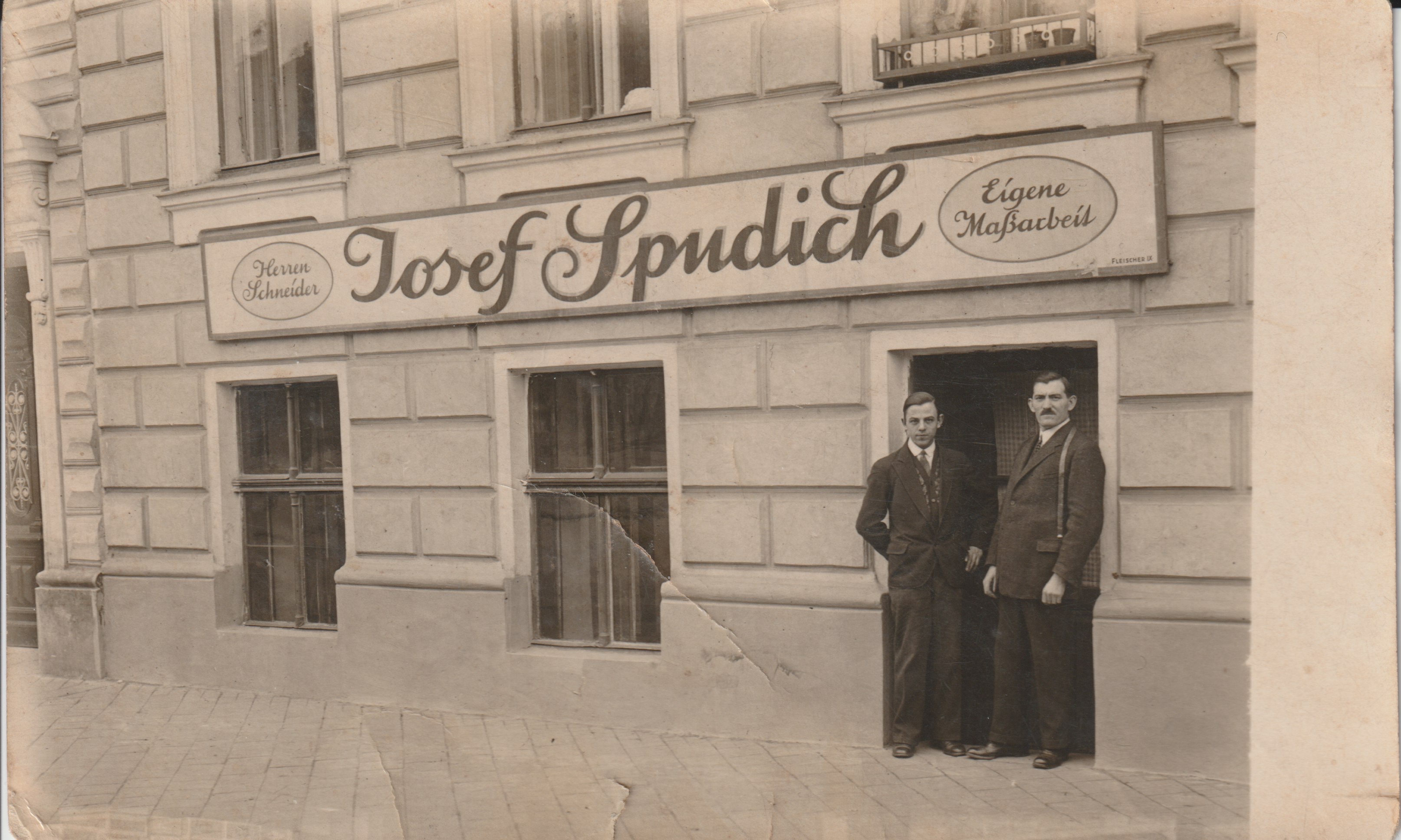 Ein altes Foto eines Geschäftsportals mit einem Schild "Herrenschneider Josef Spudich". Vor dem Geschäft stehen zwei Männer in Anzügen. Der ältere Mann rechts mit Schnurbart hat ein Maßband über der Schulter.